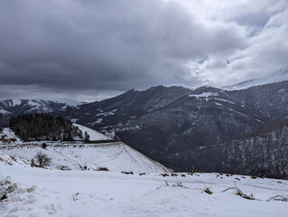 Fototapeta na wymiar Montanhas cobertas de neve num dia frio e nublado de inverno com uma pequena floresta e uma estrada a cortar a montanha