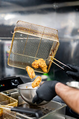 A person in black gloves transfers fried chicken from a wire basket to a metal bowl in a commercial...