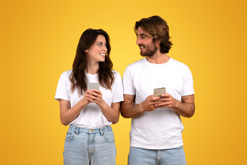 Happy couple engaging with each other, holding smartphones and smiling, woman looking at man with interest