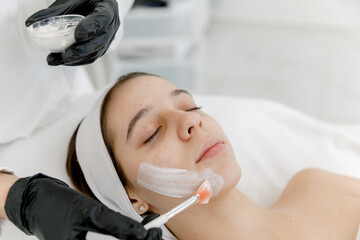 The cosmetologist applies a facial white mask to the acne woman's face.  Teen skin. Acne skin. A teenage girl at an appointment with a cosmetologist.