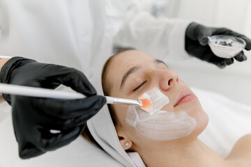 Naklejka premium The cosmetologist applies a facial white mask to the acne woman's face. Teen skin. Acne skin. A teenage girl at an appointment with a cosmetologist.