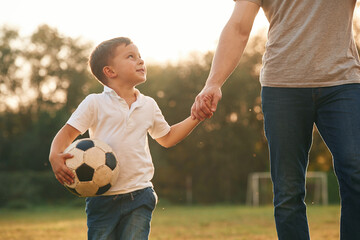 Walking and holding soccer ball. Father and little son are playing and having fun outdoors