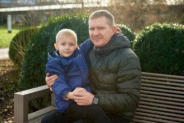 Autumnal Family Affection: Father, 40 Years Old, and Son - Beautiful 8-Year-Old Boy, Seated in the Park.