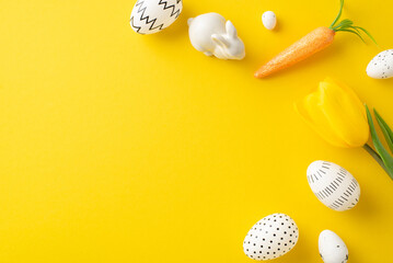 Spring festivity idea. Overhead shot of colored eggs, cute bunny figure, carrot snack for the Easter Rabbit, and tulip on a bright yellow backdrop with space for text or ads