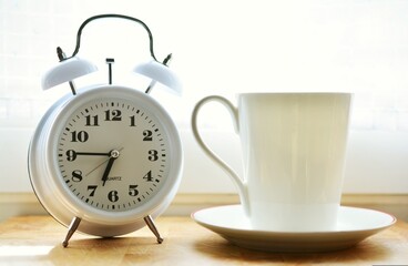 An alarm clock sits next to a steaming cup of coffee on the bedside table, signaling the start of a...