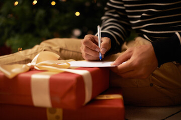 Pen, hands and man with card and present for Christmas event or party at home for family. Celebration, paper and closeup of male person writing letter with gift boxes for xmas festive holiday.