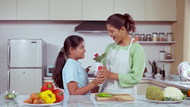 Excited daughter or kid giving flowers with greetings to mother at kitchen for mothers day celebration - concept of thankful, gratitude and childhood moments