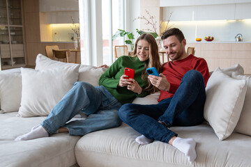 Young couple with smiles relaxing on sofa looking at screens smartphones. Happy man and woman surf...