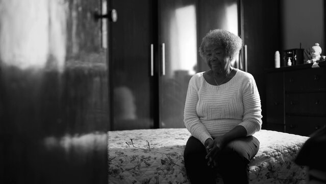 One Pensive Black Elderly Lady Seated By Bedside In Dramatic Black And White. Thoughtful Lonely Senior 80s Woman In Humble Home Solitude, Thoughtful Gaze Contemplating Life