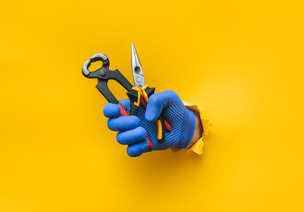 A man's hand in a blue fabric knitted glove holds wire cutters and pliers. Torn hole in yellow...