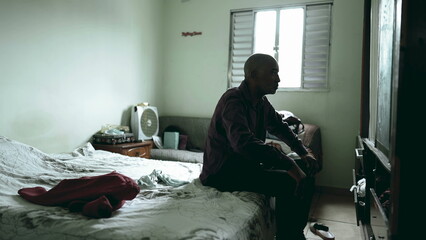 One pensive serious middle-aged man seated by bedside in a moody dark room struggling with poverty...