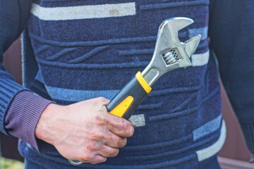 a worker in a black  sweater holds in his hand an iron manual adjustable shiny industrial wrench during the day on the street 