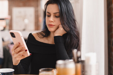 Confused puzzled brunette woman in casual clothes, sits in cafe, holds a smartphone in her hand,...