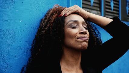 One serious black middle-aged woman regretting past mistakes leaning on blue urban wall outside...