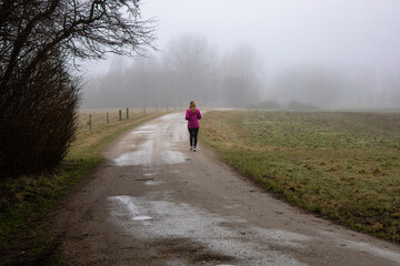 Natural landscape of a foggy morning in the countryside and an athletic woman runs along the road in sportswear. Foggy farm field.