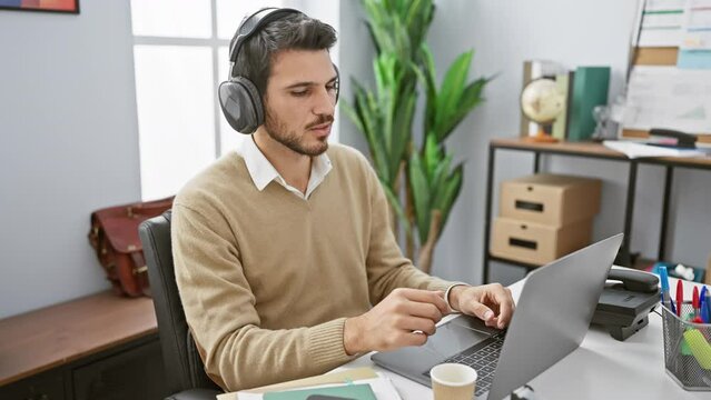 A young hispanic man with headphones works on a laptop in a modern office, depicting professionalism and concentration.