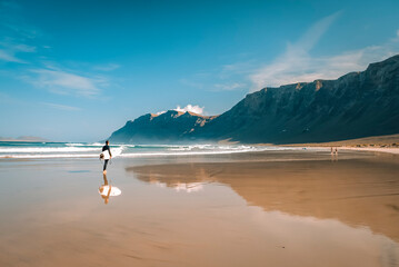 surfista si specchia su spiaggia in riva all'oceano sotto la scogliera - obrazy, fototapety, plakaty