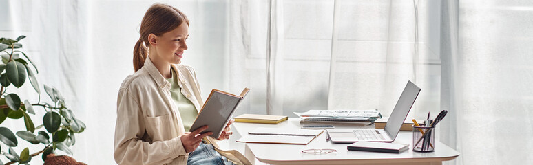 happy teenager reading book and sitting near laptop on desk, online education, horizontal banner - obrazy, fototapety, plakaty