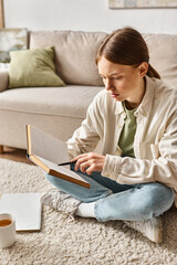 smart teenage girl reading book while doing her homework and sitting on carpet near cup of tea