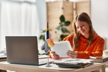 Cheerful teenage girl with tablet engaging in e-learning process near lgbtq pride flag on desk