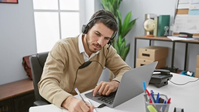 A focused hispanic man wearing a headset works on his laptop in a modern office setting.