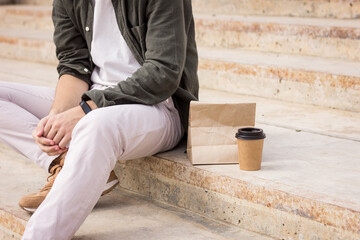 A young man sits on the steps next to a hundred coffee and food in a bag