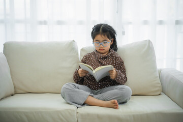 Happy asian child baby girl smiling wearing glasses and reading book while sitting on couch sofa in...