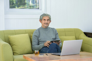 portrait asian senior man sitting on sofa in the living room,surfing on internet with tablet and laptop at home,concept of elderly people modern lifestyle with technology