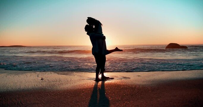 Sunset, love and couple hug at a beach with kiss, support or lifting in celebration in nature. Sunrise, romance and silhouette of people at sea excited for proposal, announcement or surprise question