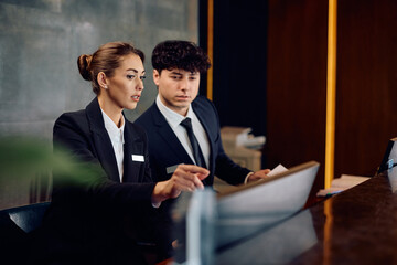 Two receptionists cooperating while working on computer at hotel front desk.