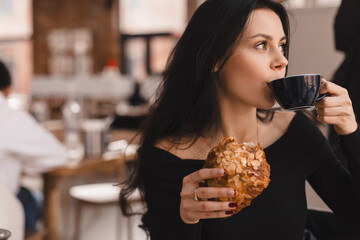 Photo of brunette teen woman ordering coffee and croissant. Young woman having breakfast with croissant and cup of coffee at the cafe. Girl drink mug of tea and hold croisant.