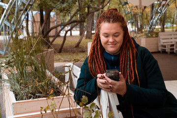 happy unusual mid adult woman with dreadlocks sits in city park using phone