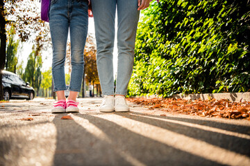 Cropped photo of two girls legs denim jeans sneakers shadow road fallen leaves green park bushes...