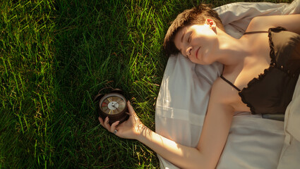 A girl wrapped in a blanket lies on green grass next to an alarm clock, promoting healthy sleep...