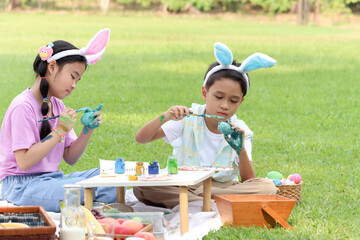 Cute Asian boy and girl with bunny ears painting egg with paintbrush while sitting together on...