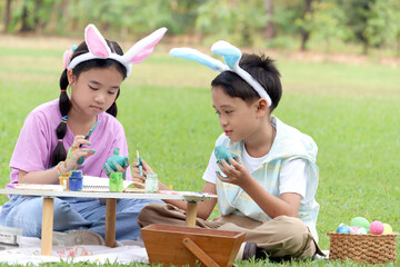 Cute Asian boy and girl with bunny ears painting egg with paintbrush while sitting together on green grass meadow in garden. Kids celebrating Easter holiday outdoor. Happy children in park on Easter