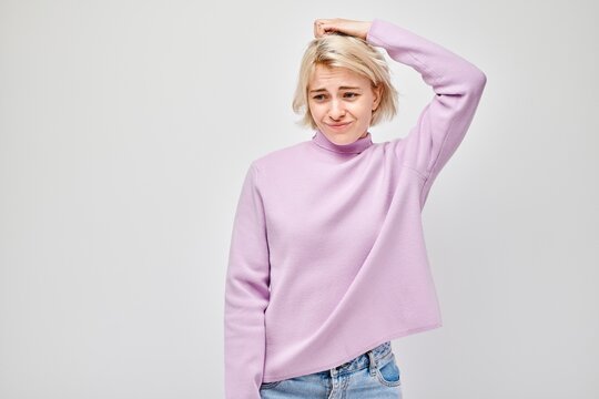 Confused young woman scratching her head, wearing a pink turtleneck on a light background.