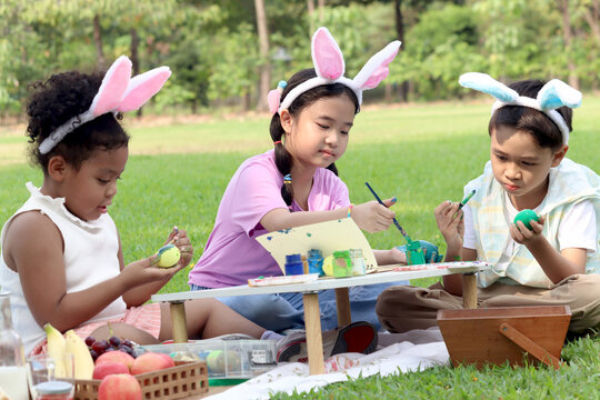 Happy three children in park, cute Asian girl and boy with curly hair African friend wear bunny ears, paint egg with paintbrush together on green grass in garden. Kids celebrate Easter holiday outdoor