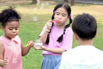 Obraz premium Happy cute Asian girl with curly hair African and friend blowing bubbles in green nature garden. Kids spending time outdoor together in meadow. Cute children having fun with bubble in summer park.