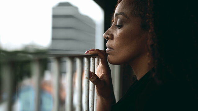 Profile close-up of a serious black woman leaning ln metal bar by home balcony gazing downwards with melancholic expression struggling but observing surroundings