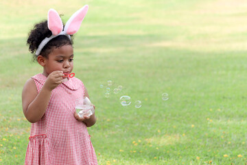 Happy cute African girl with black curly hair wearing bunny ears blowing soap bubbles in green...