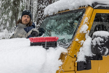 A man is pushing snow off the top of a a gold jeep with a brush.