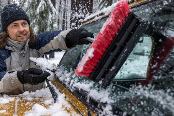 A man is brushing snow off of a car window. The man is smiling and he is enjoying the task