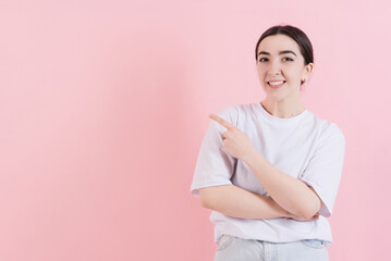 Fototapeta premium Attractive Caucasian young brunette woman in casual clothing points fingers at empty copy space for advertisement isolated on pink studio background.