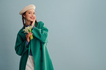 Fashionable happy smiling woman wearing trendy spring outfit with white beret, green coat, holding  bouquet of snowdrops, posing on blue background. Studio portrait. Copy, empty, blank space for text