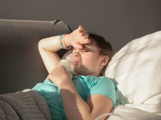 A sick little girl inhales with a nebulizer to reduce cough, lying on the couch at home, the child...