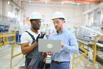 Two Diverse Professional Heavy Industry Engineers Wearing Safety Uniform and Hard Hats Working on Laptop