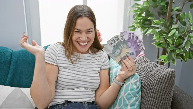 Joyful young woman with blue eyes, celebrating a triumphant win at home! huge savings with philippine peso banknotes, smiling with a winner's confidence and achievement.