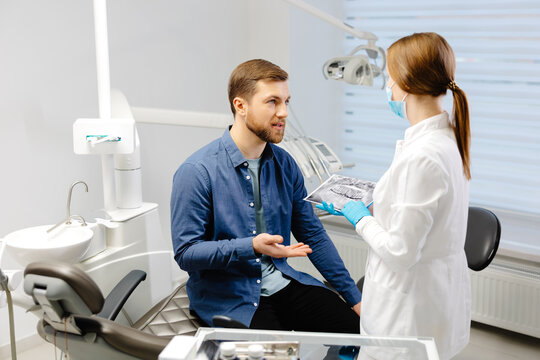 Young attractive man visiting dentist, sitting in dental chair at modern light clinic. Young woman dentist holding x ray image.