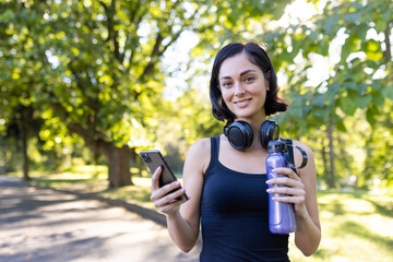 Portrait of a young female athlete and trainer standing in the park wearing headphones, holding a phone and a bottle of water in her hands, looking at the camera with a smile